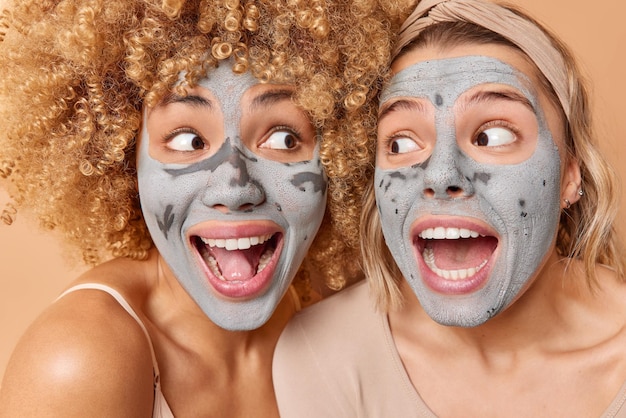 Gros plan de jeunes femmes surprises se regardent avec émerveillement garder la bouche ouverte les masques d'argile appy subissent des procédures de beauté rajeunissent la peau debout avec les épaules nues