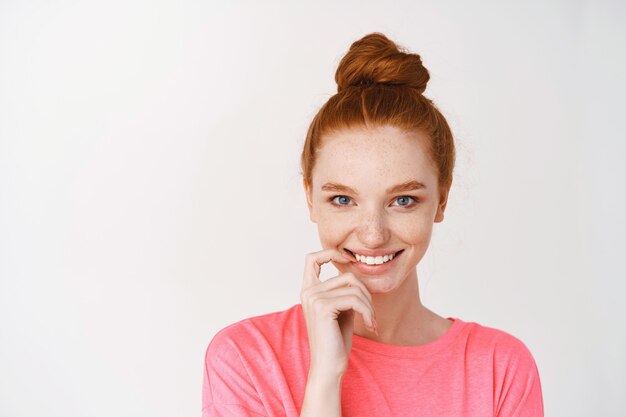 Gros plan d'une jolie adolescente aux cheveux roux peignés en chignon, souriante avec des dents blanches, montrant une peau éclatante, pas de maquillage sur le visage, debout sur un mur blanc