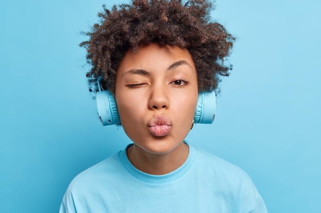 Gros plan d'une jolie femme afro-américaine affectueuse qui fait un clin d'œil, garde les lèvres arrondies et aime écouter une piste audio via des écouteurs habillés avec désinvolture isolés sur un mur bleu Concept de passe-temps