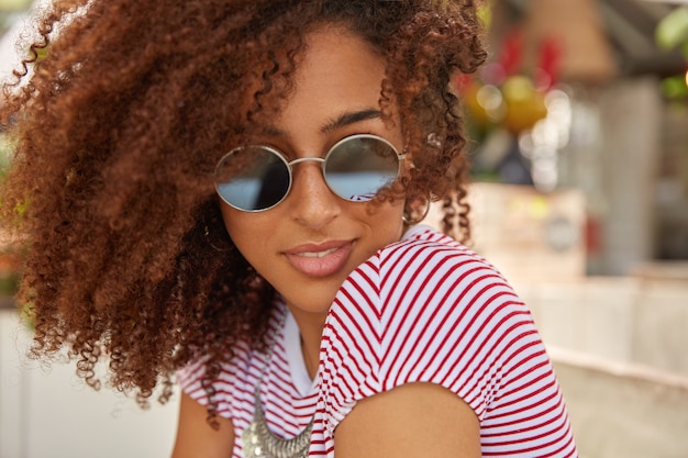 Gros plan d'une jolie femme aux cheveux bouclés, porte des nuances rondes à la mode, t-shirt décontracté, aime l'heure d'été, a son propre style, aime les vêtements à la mode