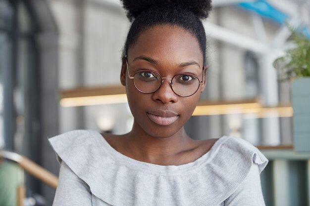 Gros plan d'une jolie femme sérieuse à la peau foncée, regarde directement la caméra avec confiance, porte des lunettes rondes, pose au bureau, a une pause après le travail.