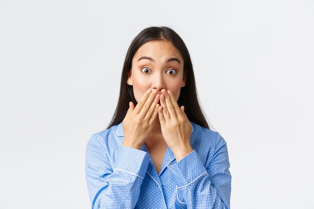 Gros plan d'une jolie fille asiatique choquée et étonnée en pyjama bleu se rendant compte de quelque chose, tenant les mains sur la bouche et les yeux émerveillés par la caméra, entend des commérages, fond blanc