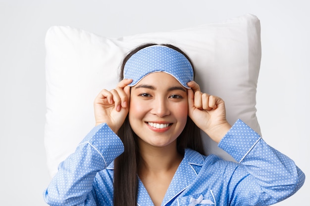 Gros plan d'une jolie fille asiatique heureuse en masque de sommeil et pyjama bleu se préparant à dormir, allongée dans son lit sur un oreiller et souriante ravie devant la caméra, posant sur fond blanc