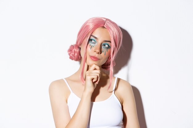 Gros plan d'une jolie fille réfléchie avec une perruque anime rose et un maquillage lumineux, regardant la bannière du coin supérieur gauche et pensant, faisant un choix, debout.