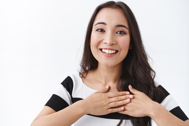 Gros plan sur une jolie et jolie jeune fille asiatique touchée, appuyez les mains sur le cœur, souriante heureuse et reconnaissante, exprimez votre gratitude ou votre plaisir de recevoir un cadeau charmant, debout sur fond blanc ravi