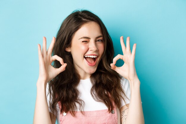 Gros plan d'une joyeuse femme caucasienne d'accord et dire oui, faire signe d'accord et faire un clin d'œil assuré à la caméra, debout satisfait sur fond bleu