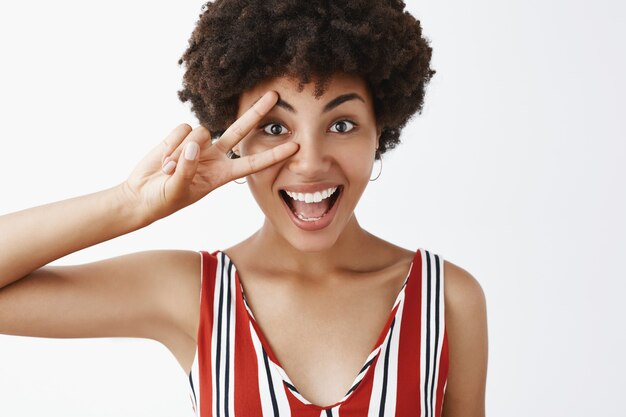 Gros plan d'une joyeuse fille disco optimiste et heureuse avec une coiffure afro en chemisier rayé montrant la victoire ou un signe de paix sur les yeux et souriant largement se sentir heureux et amusé