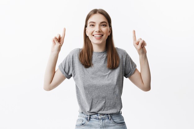 Gros plan de la joyeuse jeune belle jeune femme européenne aux cheveux noirs dans des vêtements gris décontractés souriant, avec une expression de visage détendue et heureuse, pointant à l'envers avec les deux mains.