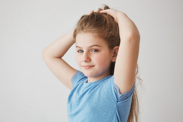 Gros plan d'une joyeuse petite fille blonde aux yeux bleus et aux joues rouges souriant, faisant ses cheveux, se préparant pour l'école