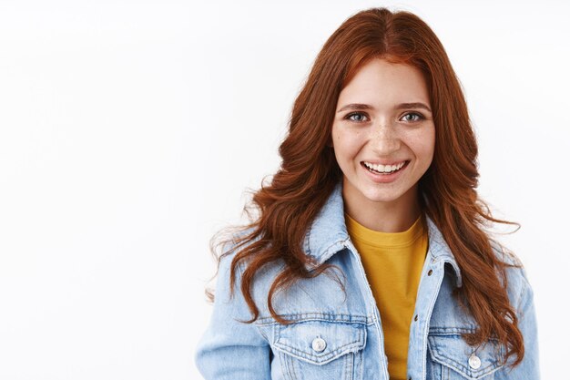 Gros plan joyeux jolie étudiante rousse avec des taches de rousseur dans une veste en jean sur un t-shirt jaune, souriant joyeusement et détendu, debout avec désinvolture sur un mur blanc aime étudier à l'étranger