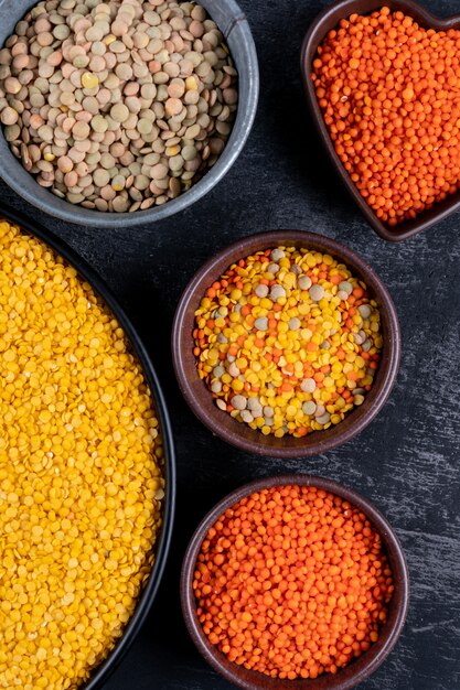 Gros plan des lentilles vertes, rouges et jaunes dans des bols de différentes couleurs et une casserole noire sur une table en pierre noire. verticale