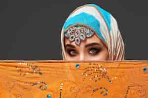 Photo gratuite gros plan d'une magnifique jeune femme avec un maquillage professionnel portant un élégant hijab coloré décoré de paillettes et de bijoux. elle couvre son visage avec un châle orange et regarde e