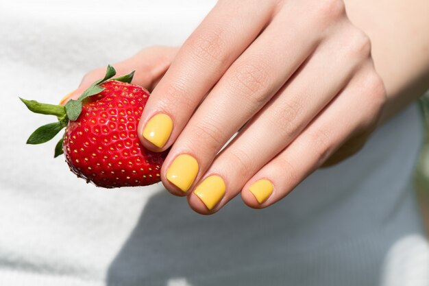 Gros plan d'une main féminine avec une manucure de conception d'ongles assez jaune tenant une fraise mûre.