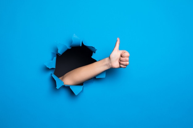 Photo gratuite gros plan de la main de la femme avec le pouce en l'air geste à travers le trou bleu dans le mur de papier.