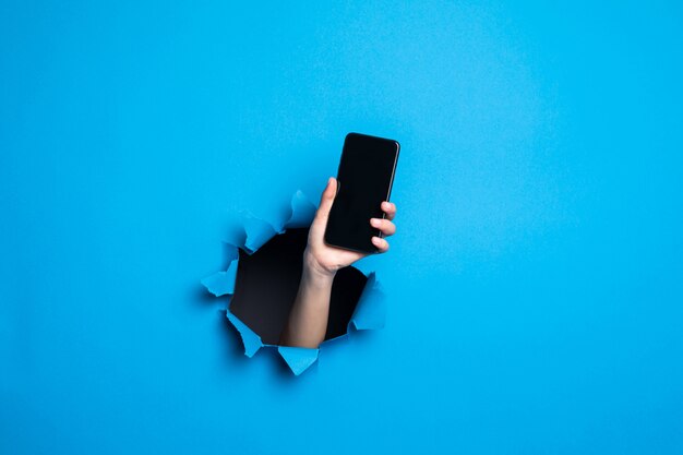 Gros plan de la main de femme tenant le téléphone avec screan pour adv à travers le trou bleu dans le mur de papier.