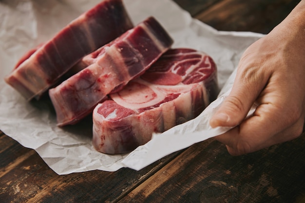 Gros plan main tient du papier kraft blanc avec trois excellents steaks de jambe avec de l'os dessus, tout sur la vieille table en bois brossé