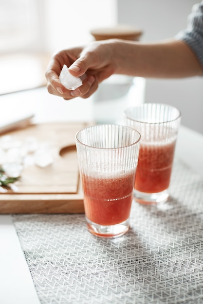 Gros plan des mains de femme mettant des morceaux de glace dans des verres avec smoothie sain de désintoxication de pamplemousse.