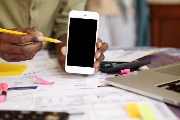 Gros plan des mains de l'homme à la peau sombre tenant un téléphone portable à écran blanc et un crayon