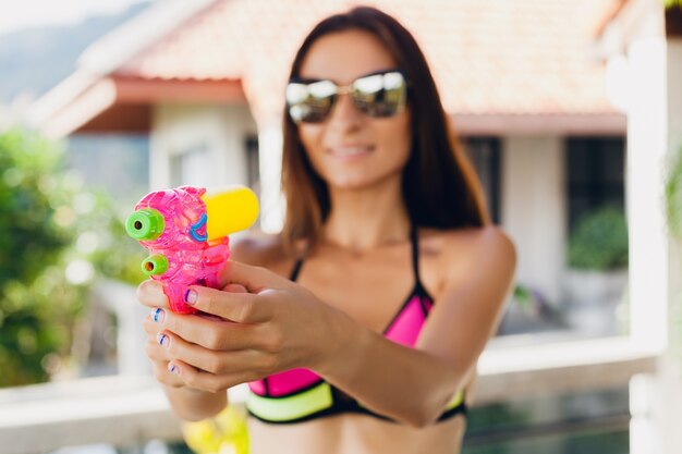 Gros plan des mains de jolie femme heureuse souriante jouant avec jouet de pistolet à eau à la piscine en vacances tropicales d'été sur la villa hôtel s'amuser en maillot de bain bikini, style coloré, ambiance de fête