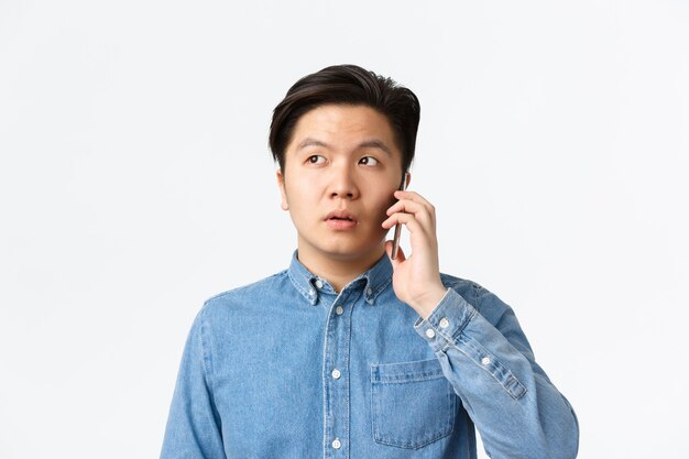 Gros plan d'un mec asiatique indécis qui s'espace pendant un appel téléphonique, discute et détourne le regard confus, debout sur fond blanc. Homme tenant un smartphone près de l'oreille, commande la livraison de nourriture.