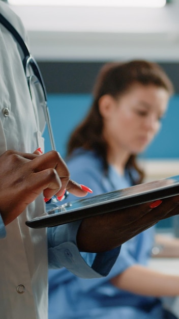 Gros plan sur un médecin afro-américain utilisant une tablette avec écran tactile pour obtenir des informations sur les soins de santé dans le cabinet. Médecin noir avec blouse blanche et stéthoscope tenant un gadget moderne pour les soins médicaux