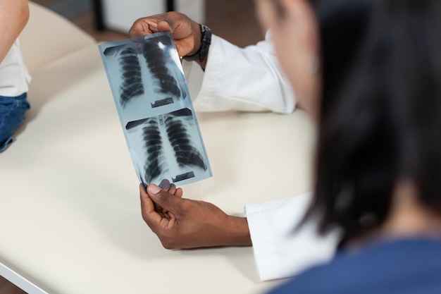 Photo gratuite gros plan d'un médecin pédiatre afro-américain tenant une radiographie pulmonaire à la main discutant des résultats médicaux lors de la consultation au bureau de l'hôpital. homme thérapeute analysant la radiographie. service de médecine