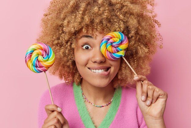 Gros plan d'un modèle féminin aux cheveux bouclés joyeux tenant deux délicieuses sucettes multicolores mord les lèvres s'amuse ne se soucie pas des effets nocifs du sucre isolé sur fond rose