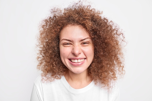 Gros plan d'un modèle féminin européen sincère et heureux qui s'amuse avec des sourires narquois face à la joie a des cheveux bouclés et touffus habillés d'un t-shirt décontracté souriant positivement isolé sur un mur blanc. Notion d'émotions