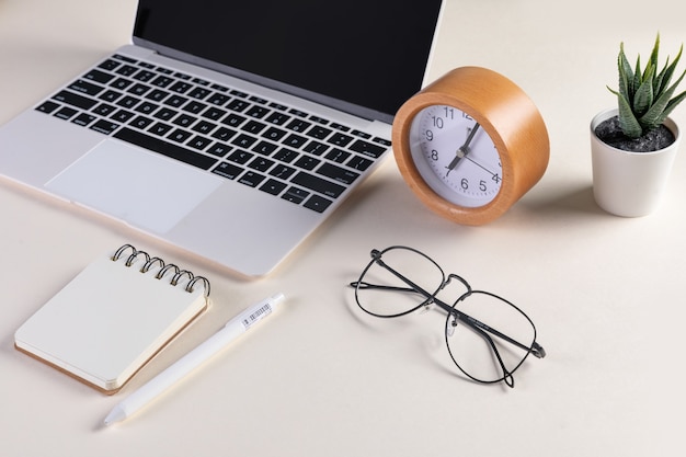 Gros plan sur un ordinateur portable ouvert, des lunettes, un bloc-notes, un stylo, une horloge et une plante