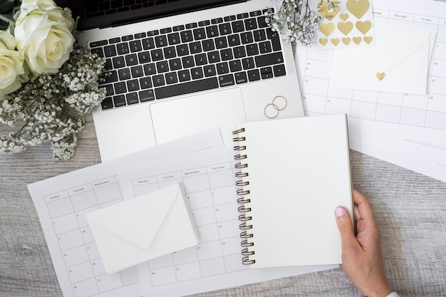 Gros plan, personne, tenue, blanc, cahier spirale, à, ordinateur portable; anneaux de mariage; fleur; enveloppe et calendriers sur un bureau en bois