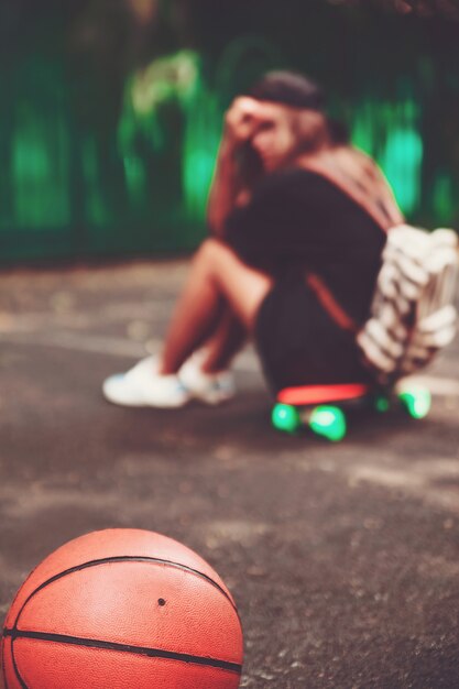 Gros plan photo ballon de basket avec une fille assise sur un shortboard en plastique orange penny sur l'asphalte
