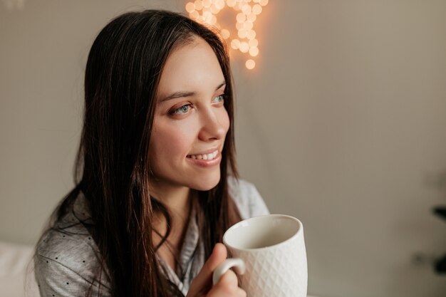 Gros plan photo d'intérieur de femme heureuse souriante aux cheveux noirs regardant la fenêtre tout en buvant du café.