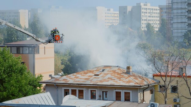 Gros plan sur des pompiers sur un camion à plate-forme aidant à lutter contre l'incendie d'un bâtiment en feu. Pompiers avec équipement et eau travaillant pour éteindre les flammes sur le toit de la maison dans le paysage de la ville.