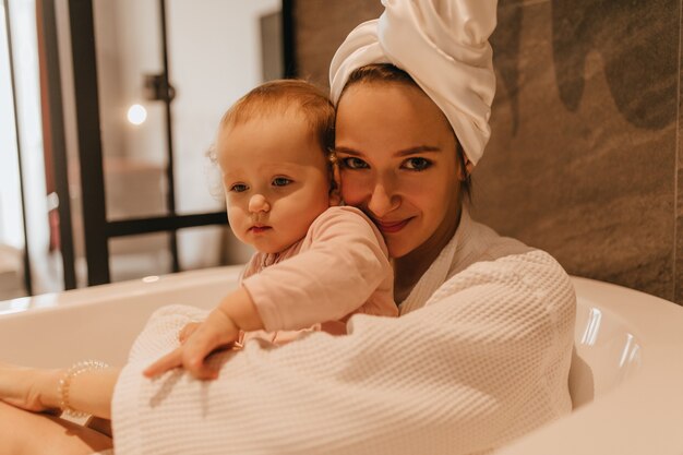 Gros plan portrait de dame en robe blanche et serviette sur sa tête assise avec sa fille dans un bain profond blanc.