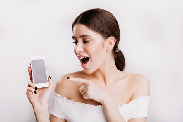 Gros plan portrait de femme brune souriante en haut blanc, regardant le téléphone avec étonnement.