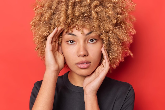 Gros plan portrait d'une femme sérieuse confiante avec des cheveux blonds bouclés touche le visage regarde directement la caméra porte un t-shirt noir décontracté isolé sur fond rouge montre la beauté naturelle sans maquillage