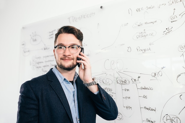 Gros plan portrait de jeune homme aux cheveux noirs debout près du tableau blanc et parler au téléphone au bureau. Il porte une chemise bleue, une veste sombre, des lunettes. Il sourit sur le côté.