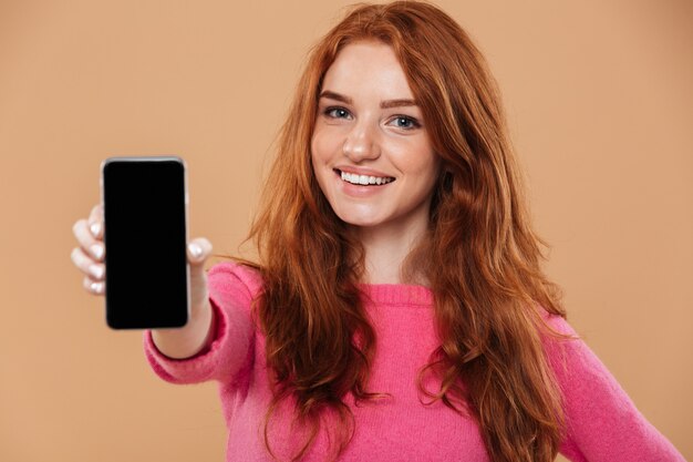Gros plan le portrait d'une jolie rousse souriante montrant un smartphone