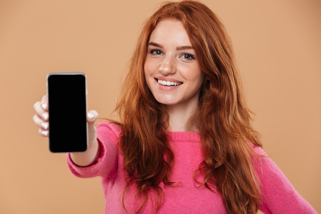 Gros plan le portrait d'une jolie rousse souriante montrant un smartphone