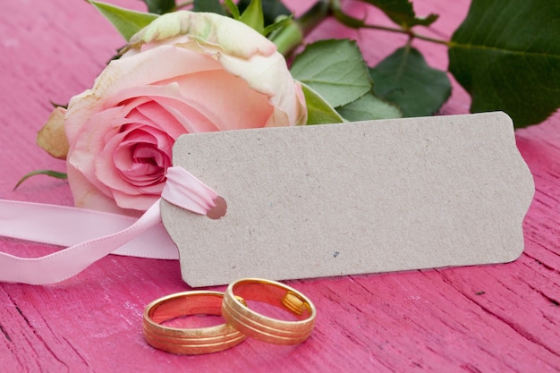 Gros plan d'une rose rose, une balise avec un espace pour le texte et deux anneaux de mariage en or sur une table rose