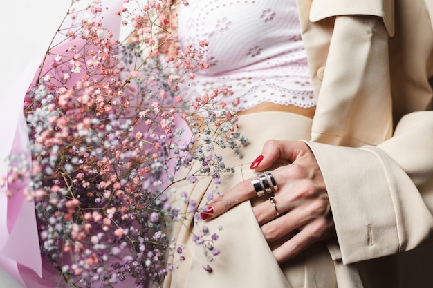 Gros plan sans tête femme en costume beige et soutien-gorge blanc tenir bouquet de fleurs séchées colorées manucure rouge deux anneaux sur les doigts