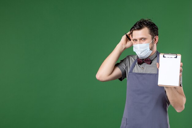 Gros plan d'un serveur masculin confus en uniforme avec un masque médical et tenant un carnet de commandes sur fond vert