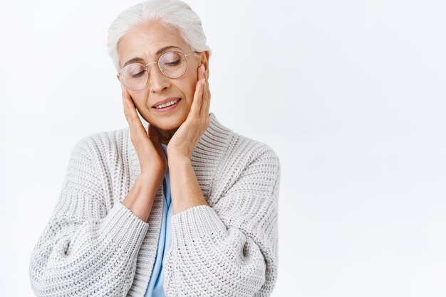 Gros plan tendre romantique et mignonne grand-mère femme senior aux cheveux gris peignés dans des verres touchant le visage avec les yeux fermés souriant joyeusement en prenant soin des rides appliquer une crème anti-âge