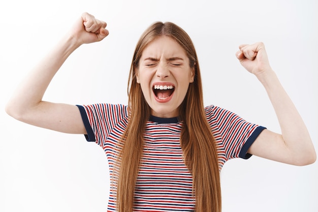 Photo gratuite gros plan triomphant, joyeusement jeune femme en t-shirt rayé, levant les mains en criant, enracinant pour l'équipe préférée, fermez les yeux et la pompe à poing, célébrant la victoire, debout sur fond blanc joyeux