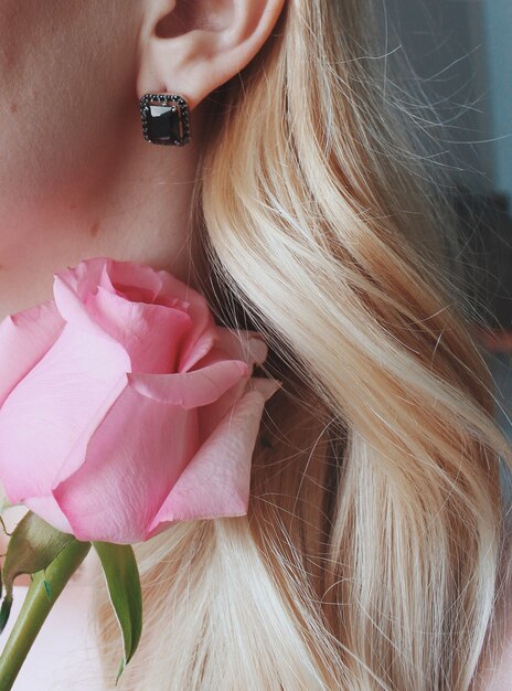 Gros plan vertical tiré d'une femme blonde portant une boucle d'oreille avec une perle noire tenant une rose rose