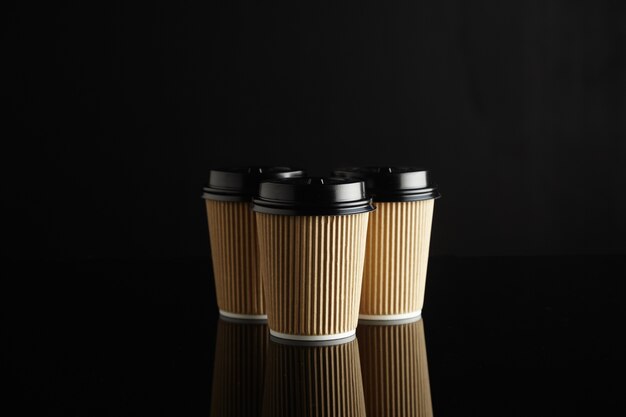 Un groupe de 3 tasses à café jetables en carton ondulé marron clair identiques avec couvercles noirs au milieu d'une table réfléchie noire avec un mur noir derrière.