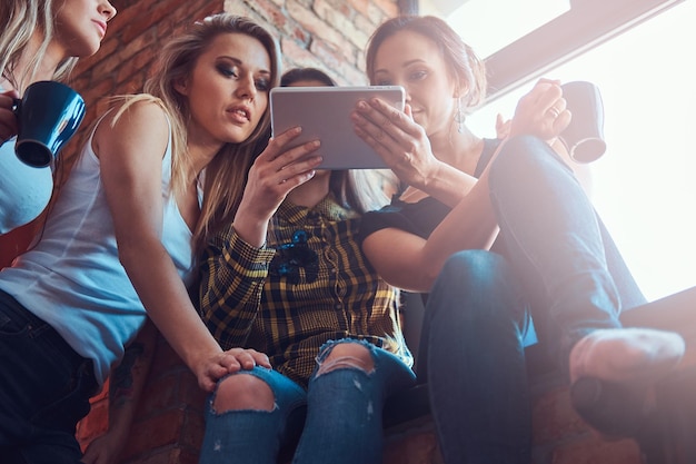 Groupe d'amies en vêtements décontractés discutant tout en regardant quelque chose sur une tablette numérique dans une pièce avec un intérieur loft.