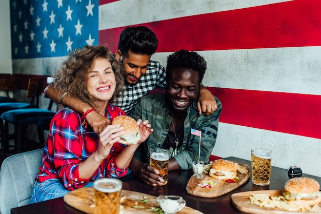 Groupe d'amis attirants s'embrassant, mangeant des hamburgers, parlant et souriant tout en passant du temps ensemble dans un pub gastronomique.