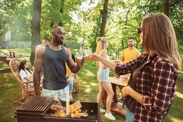 Groupe d'amis heureux ayant de la bière et un barbecue en journée ensoleillée. Repos ensemble en plein air dans une clairière ou une cour. Célébrer et se détendre, rire. Mode de vie d'été, concept d'amitié.