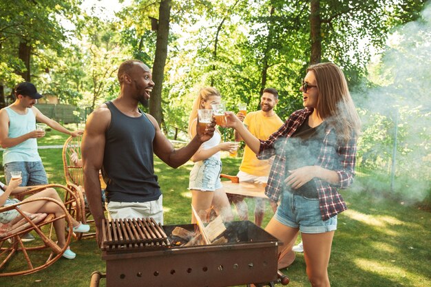 Groupe d'amis heureux ayant de la bière et un barbecue en journée ensoleillée. Repos ensemble en plein air dans une clairière ou une cour